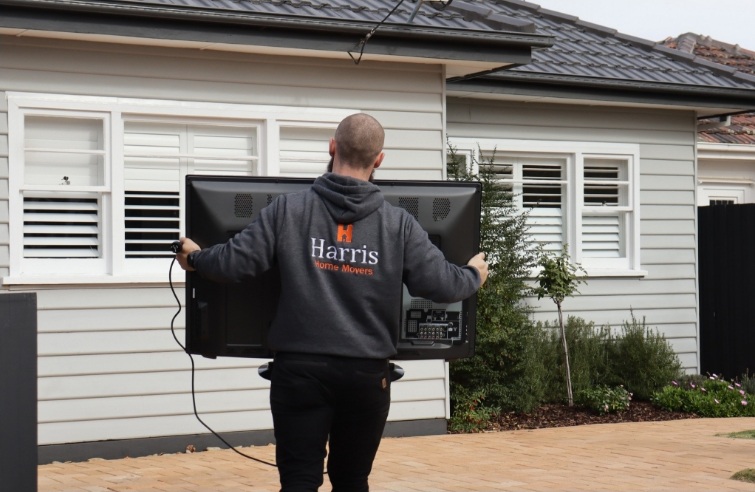 A member of the Harris Movers team carrying a tv into a house wearing a branded hoodie.
